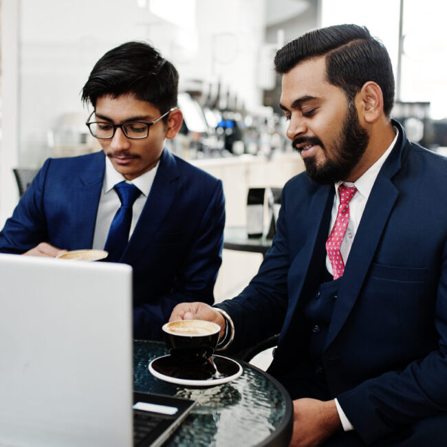 Two indian business man in suits sitting at office on cafe, look Two indian business man in suits sitting at office on cafe, looking at laptop and drinking coffee.